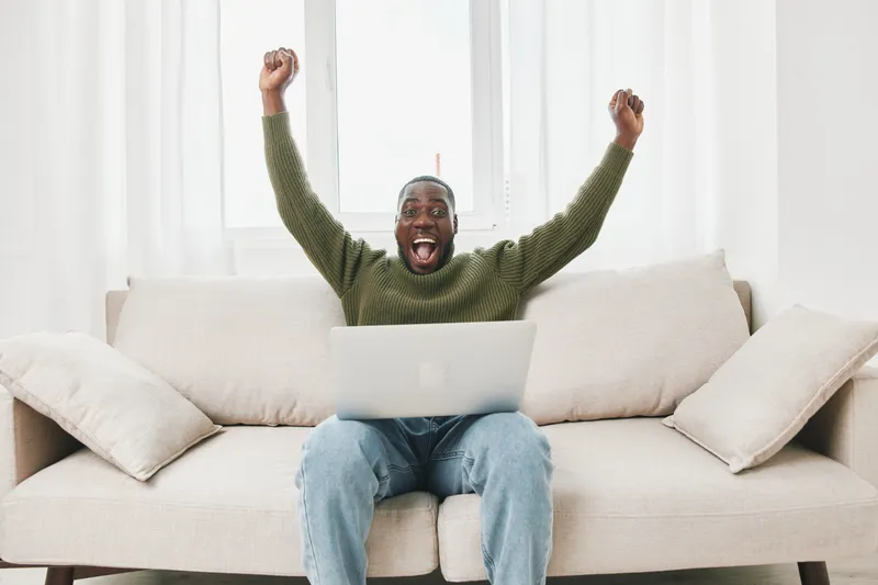 Excited young Black man celebrating success while working on a laptop in a cozy living room Happiness, motivation, and productivity are emphasized