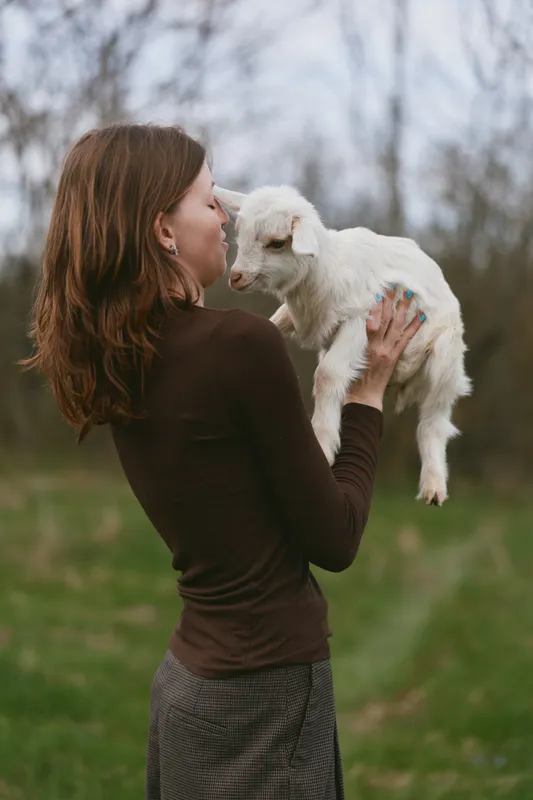 Cheerful woman outdoors, holding a goat kid close, sharing a gentle kiss, sunlit meadow scene, casual attire, tender moment, rustic vibe, soft natural colors, peaceful connection between human and