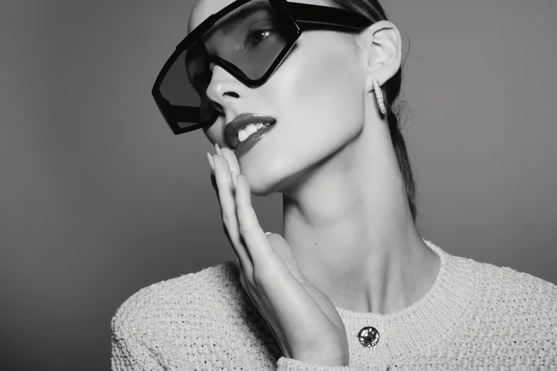 Stylish Caucasian woman with pale skin wearing large sunglasses and a textured sweater poses with hand on cheek in monochrome studio portrait.
