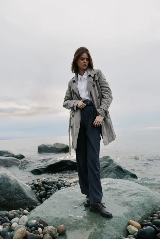 Young woman in gray coat and dark pants stands on rocky shore near calm sea under cloudy sky. Casual fashion and outdoor lifestyle concept with natural muted colors and moody atmosphere.