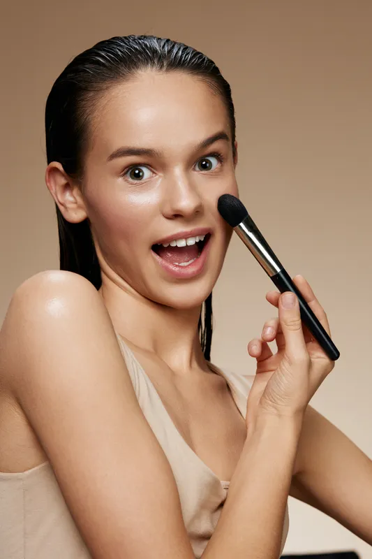 Young woman applying makeup with a brush, expressing joy and confidence, highlighted by a neutral background and soft tones Perfect for beauty and skincare themes