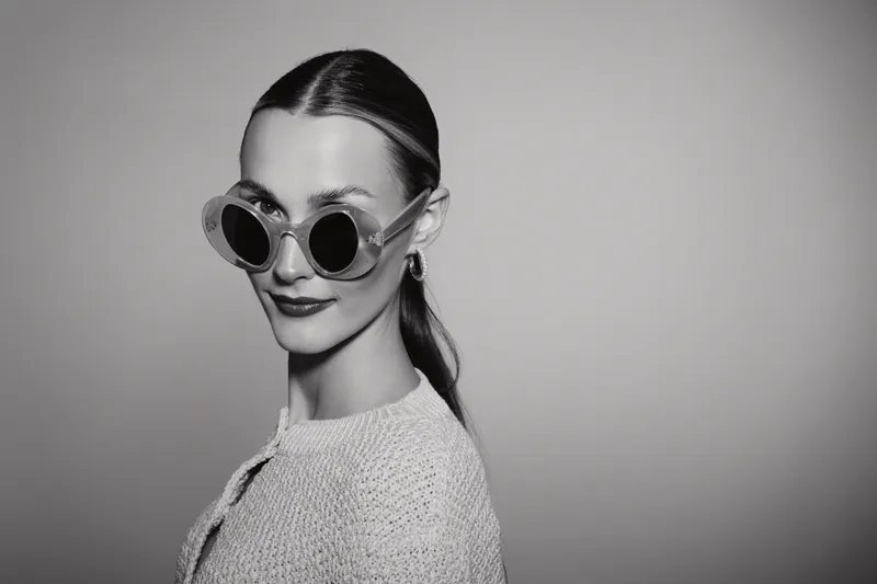 Portrait of a young white woman with pale skin wearing large round sunglasses and a knitted sweater. Studio black and white photo with neutral background and confident mood.