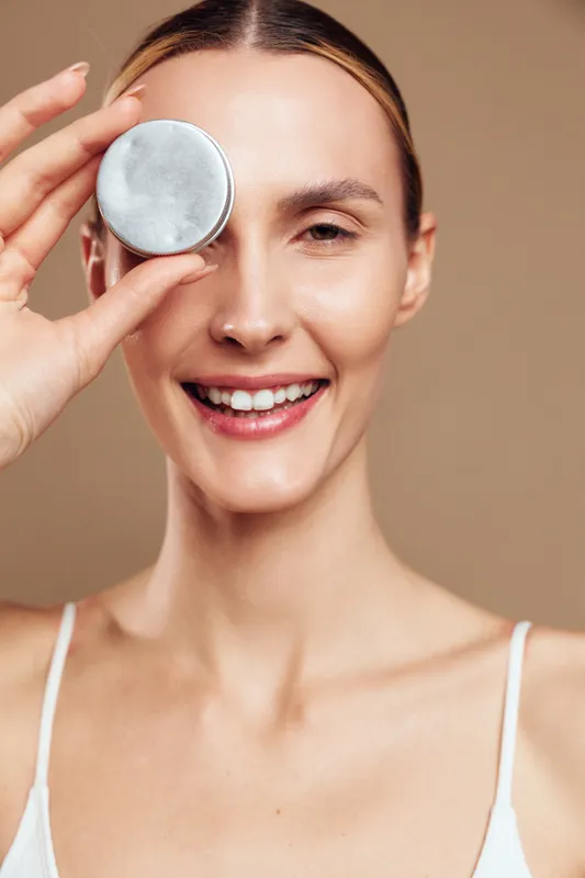 Cheerful woman closeup, smiling with a round pad held to her eye, soft lighting, natural skin, skincare routine moment, light background, relaxed pose, fresh appearance
