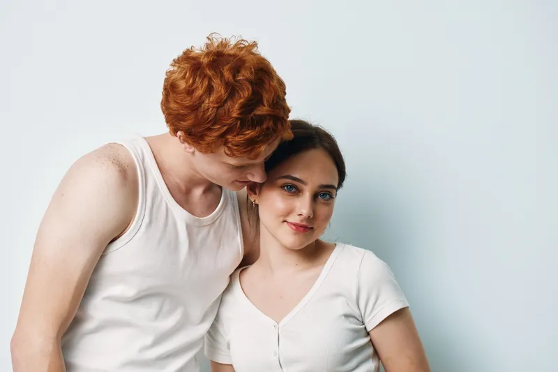 Young couple standing close with affectionate pose, redhead man wearing white tank top and brunette woman in white shirt smiling gently against plain wall background.
