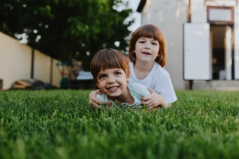 Two joyful children playing on green grass, one smiling boy enjoying the moment. A fun and carefree atmosphere in a backyard setting.