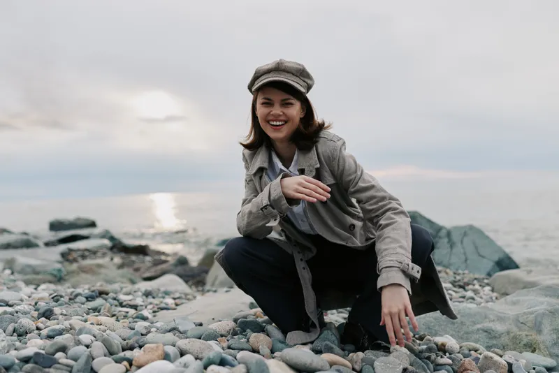Smiling young woman in casual clothes and hat crouches on rocky beach near calm sea. Overcast sky and soft natural light create peaceful mood for outdoor lifestyle and travel concept.