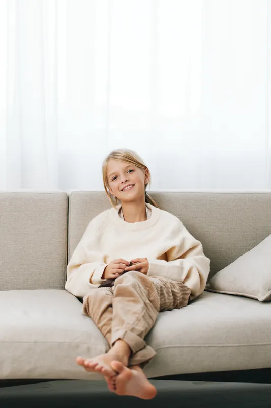 Young girl sits barefoot on a light beige sofa, smiling softly at the camera. Cozy casual outfit, warm natural light, relaxed pose, inviting scene of home comfort and everyday joy.