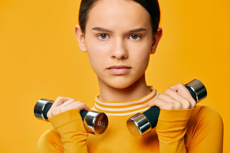 Fitness portrait of a woman in an orange top holding dumbbells, ready for a workout. Strong, focused and energetic against a vibrant background highlighting training and health.