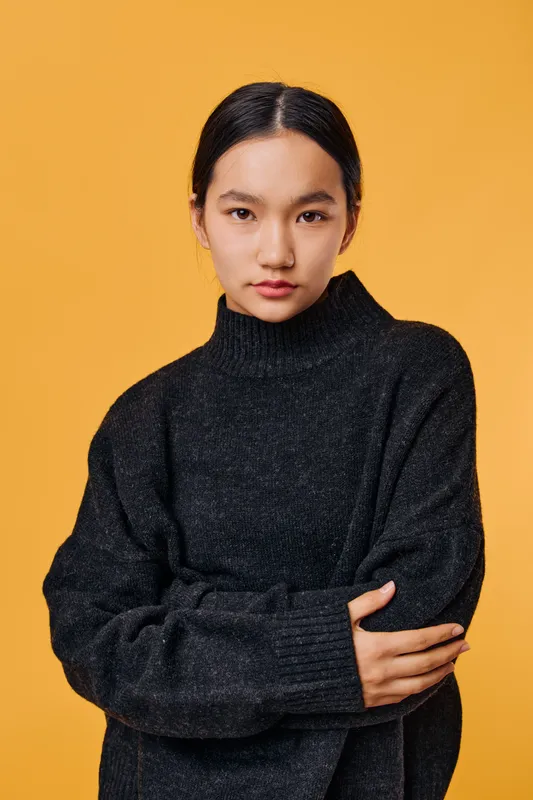 Young woman with dark hair and serious expression wearing black sweater standing with crossed arms on bright yellow background studio portrait. Female model, casual clothing and minimalism.