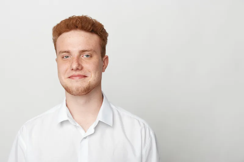 Young man with red hair and beard wearing white shirt smiles gently against plain light background. Portrait of confident male with casual style and natural expression.