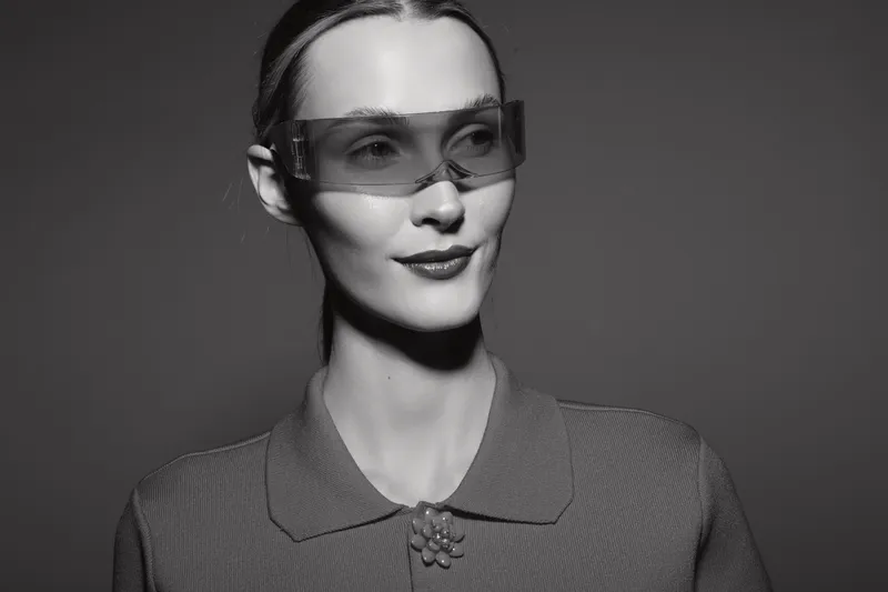 Portrait of a young white woman with pale skin wearing futuristic sunglasses and a textured collared shirt. The black and white photo highlights modern fashion and minimalism.