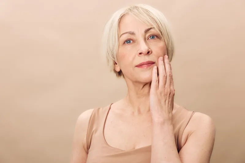 Mature woman with short blonde hair touches her face gently in a neutral beige background. Concept of natural aging, skincare, beauty, and self-confidence in middle age.