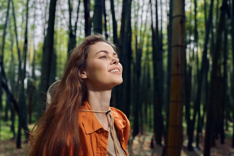 Woman portrait in forest nature smiling outdoors among tall trees with redhair and orange jacket, enjoying fresh air, peaceful woodland scenery and light filtering through leaves.