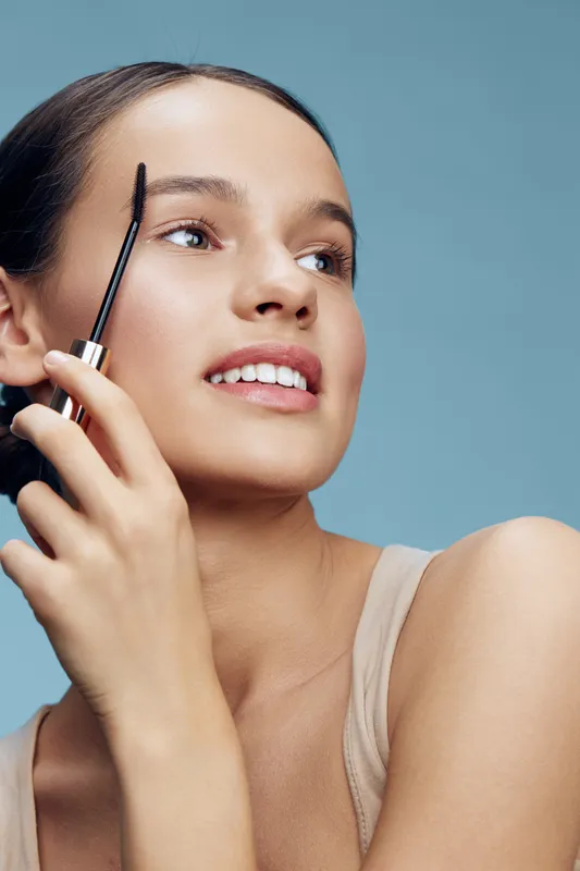 Young woman applying mascara with a joyful expression against a soft blue background, showcasing beauty and cosmetics, perfect for skincare and makeup themes