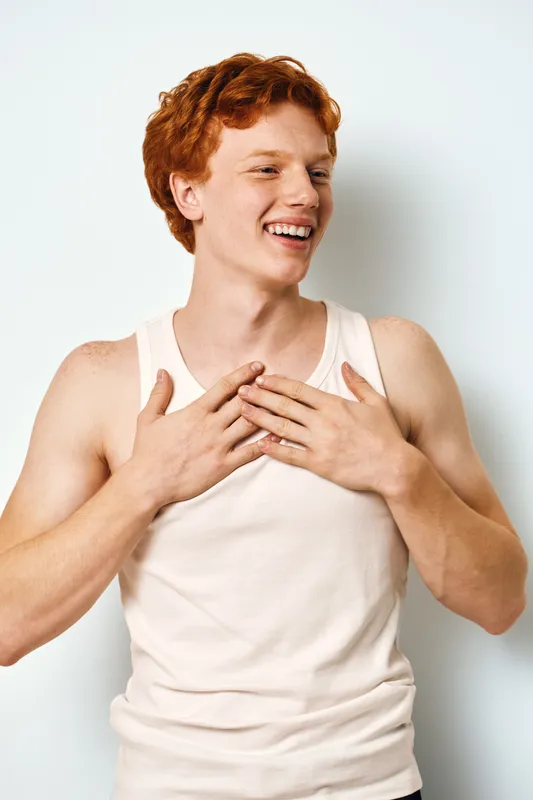 Young man with red hair in white sleeveless shirt smiling and holding hands on chest isolated on white background. Casual expression and happy mood.