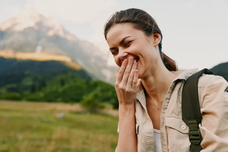 A happy young woman with a backpack laughs covering her mouth in a green mountain landscape during daytime. Lifestyle concept of travel, joy, and nature adventure.