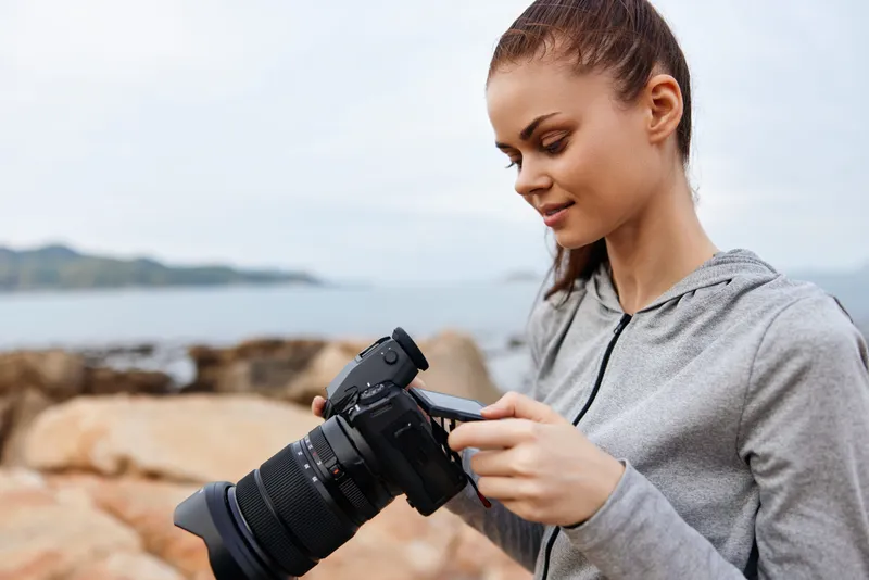Young woman capturing scenic views with a professional camera, wearing a casual gray hoodie, smiling, amidst rocky beach background showcasing serene colors