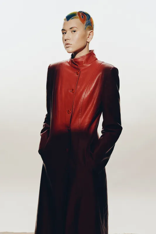 woman with colorful short hair wearing a long red leather coat stands confidently with hands in pockets, minimalist white background, fashion style
