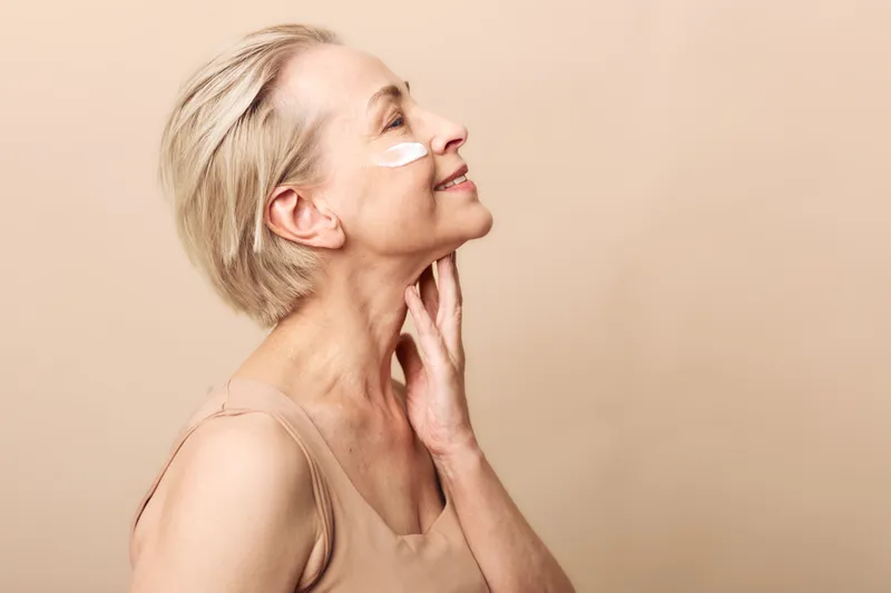Smiling mature woman with short hair applies white face cream on cheek, eyes closed, touching neck. Concept of skincare, aging care, beauty routine, and natural wellness in studio light.