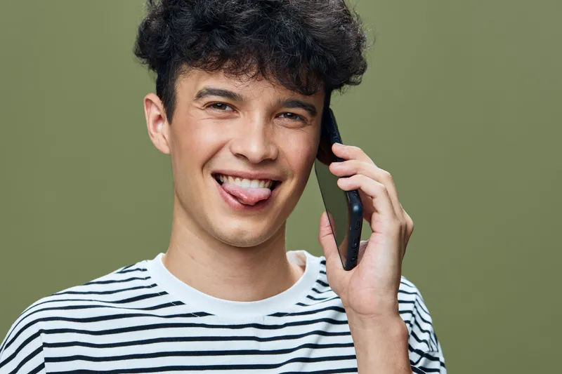 young man talking on smartphone with playful expression wearing striped shirt in front of olive green background, casual lifestyle, communication concept