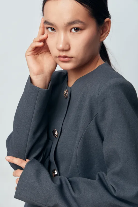 Young woman with black hair and natural makeup wearing a gray buttoned jacket posing with hand near face on light gray background. Serious model with stylish outfit and clean look.