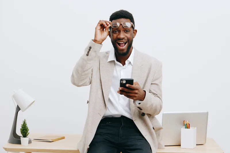 Smiling happy young man with dark skin, glasses, casual beige jacket, and white shirt holding smartphone in office environment, isolated on white background. People lifestyle concept