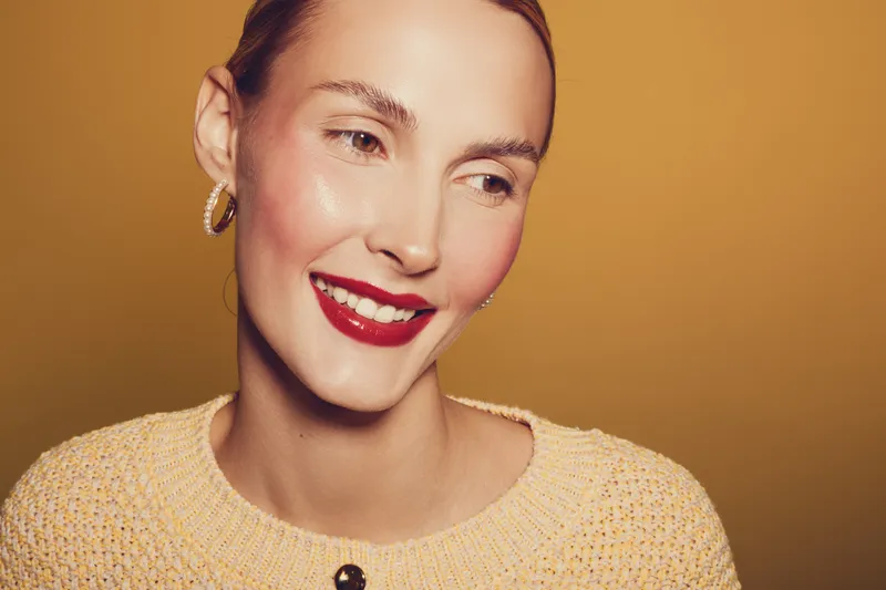 Smiling woman with red lipstick and hoop earrings looks away on yellow background. Close-up portrait with natural skin and warm lighting. Concept of beauty, fashion, and happiness.