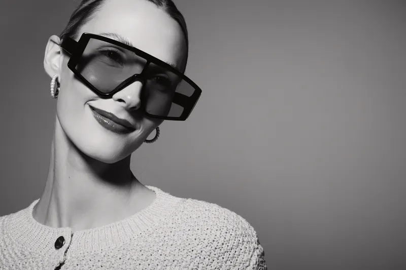 Stylish Caucasian woman with pale skin wearing oversized black sunglasses and white sweater smiles confidently on grey background.