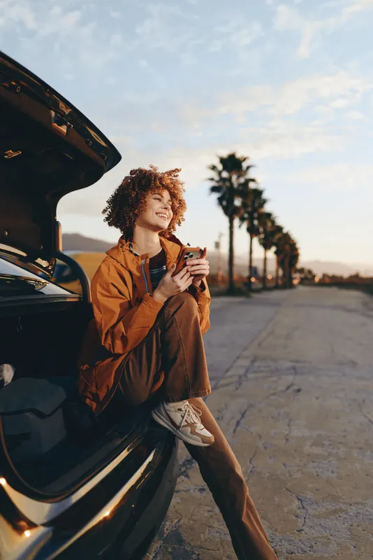 Curly haired woman at an open car trunk holding a coffee cup outdoors at sunset, travel lifestyle moment, road perspective, warm natural light, relaxed expression and casual outfit for daily life