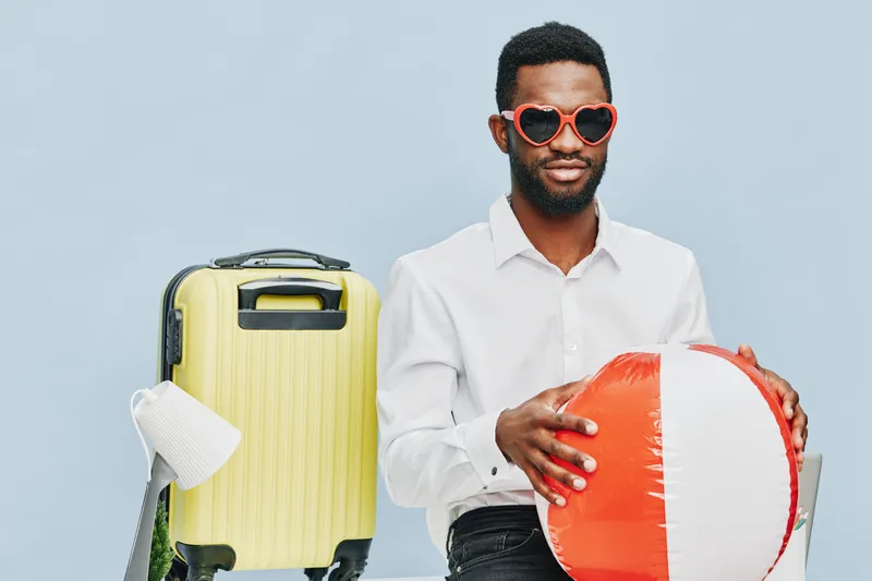 Man holding inflatable ball with luggage in background on light blue studio backdrop, casual attire, cheerful expression, travel, vacation, leisure, summer atmosphere, holiday concept, lifestyle photo