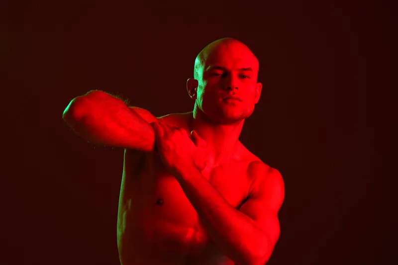 Intense man striking a pose in dramatic lighting, showcasing strong muscles and artistic expression, emphasizing movement and emotion in a vibrant atmosphere.