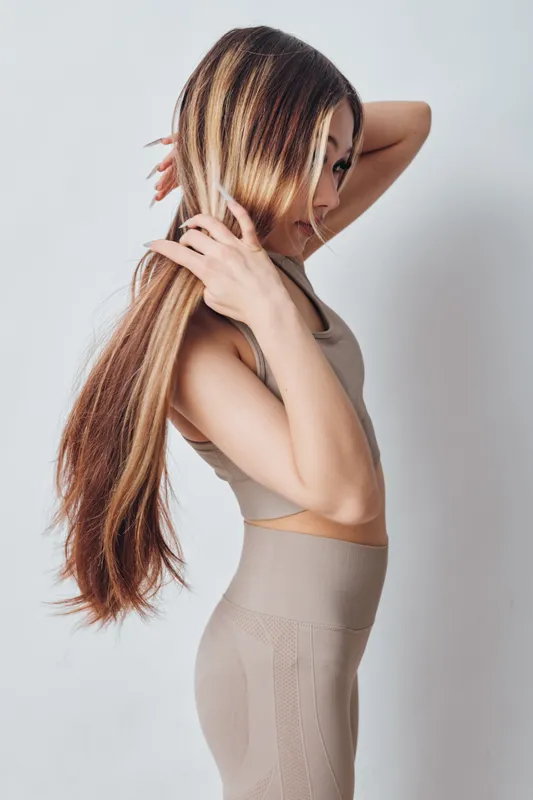 Young woman with long straight hair wearing beige fitness outfit standing sideways on white background. She is touching her hair with hands showing relaxed and calm mood in studio.