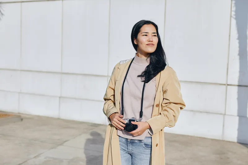 Woman wearing a beige coat stands outdoors with a confident pose, casual style on a sunlit street, emphasizing texture, mood, and relaxed fashion in a minimal, modern shot