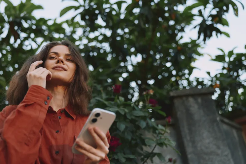 Smiling woman with smartphone outdoors in a garden, casual attire, natural light, lifestyle moment emphasizing self expression and everyday leisure
