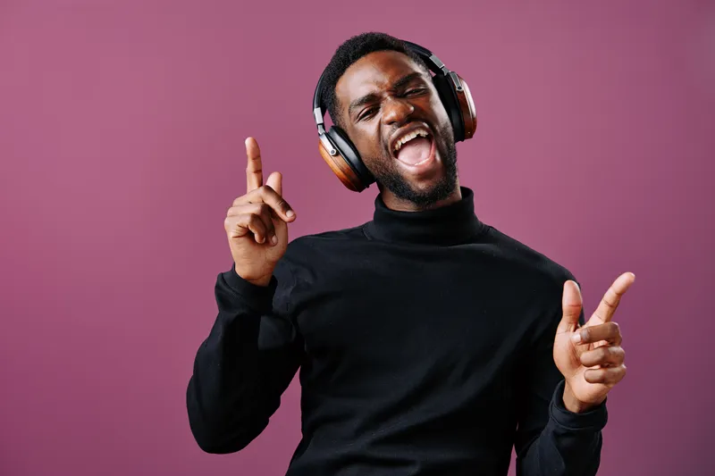 Happy young Black man enjoying music with headphones, dancing joyfully against a pink background, showcasing lively emotions and stylish clothing