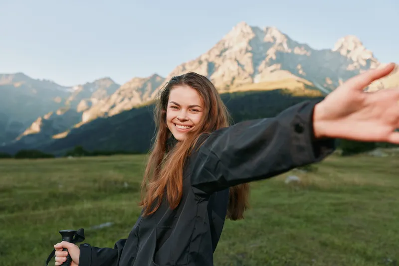 A cheerful young woman stands outdoors in a grassy field with majestic mountains in the background, smiling warmly as she extends an arm toward the camera, creating a lively, adventurous lifestyle