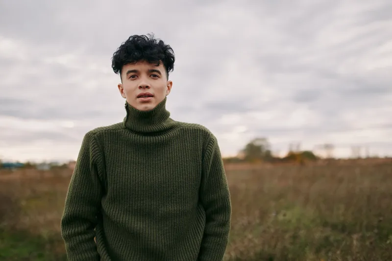 A candid portrait of a young man in a thick green turtleneck standing in a vast field, conveying authenticity and credibility through simple, natural presence.