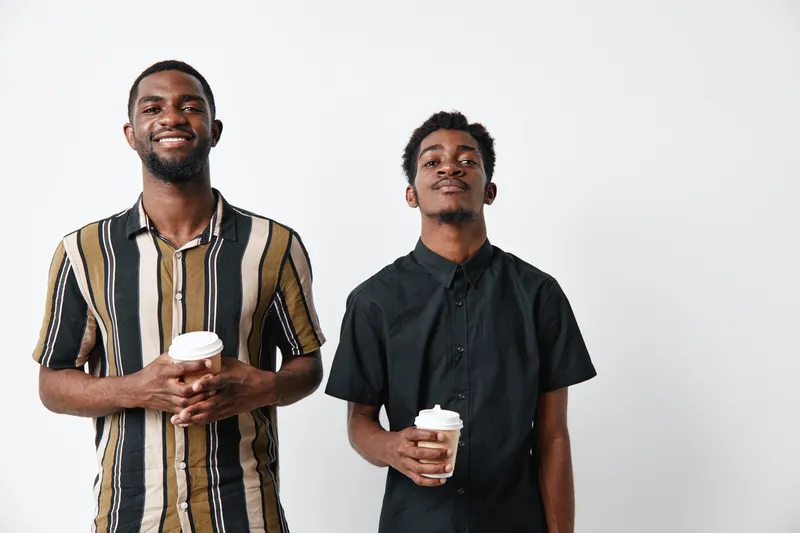 Two young black men with dark skin holding coffee cups standing against white background smiling and looking at camera in casual and formal shirts indoors.
