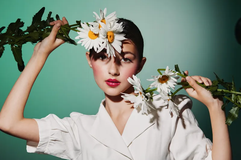 Young woman in a white blouse holds white daisies framing her face. Creative beauty and fashion styling with a soft teal background and dramatic shadows.