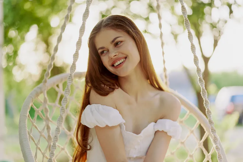 Young woman smiling in a white dress, sitting on a swing in a sunny park with blurred green trees in the background, evoking a feeling of joy and tranquility