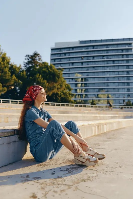 young woman sitting outdoors in casual denim outfit and red bandana enjoying sunny day with modern building and trees in background