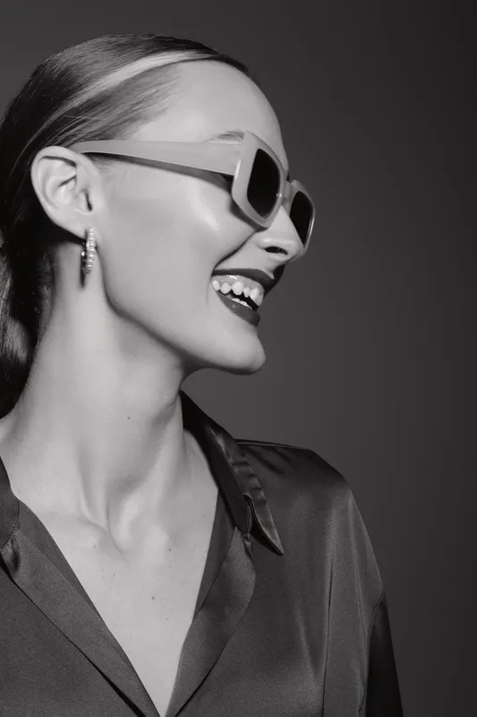 Young Caucasian woman with fair skin smiling joyfully wearing sunglasses and earrings against dark background. Concept for fashion, style, happiness, and beauty portrait.