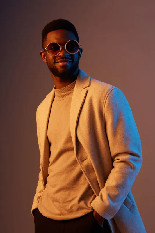 Stylish Black man in a beige coat and round sunglasses, showcasing a confident smile against a colorful gradient background, representing modern fashion