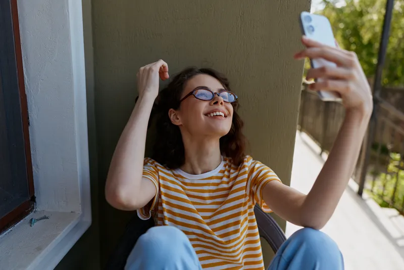 Young woman on a balcony takes a cheerful selfie, wearing a striped t shirt, glasses, relaxed smile, home lifestyle mood enhanced by natural light and casual moment.