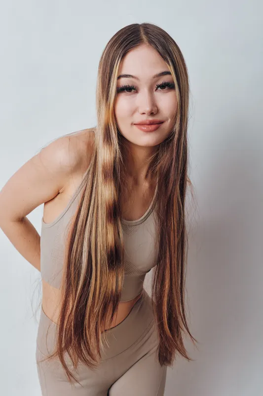 Young woman with long straight hair posing in beige workout clothes against white background, looking confident with natural makeup and relaxed expression.