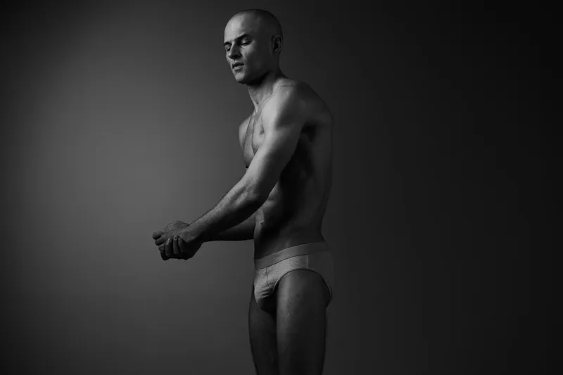 Black and white image of a man posing confidently, showcasing his physique with a minimalist background that enhances his strong silhouette.