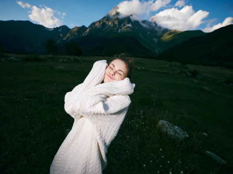 Softly smiling woman in a thick white sweater hugs herself in a green meadow, distant mountains and a bright blue sky forming a peaceful outdoor scene, conveying warmth, comfort, and connection with