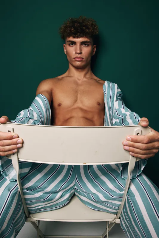 Young man with curly hair sitting on chair backwards wearing striped pants and open shirt showing bare chest against dark green background in studio portrait.