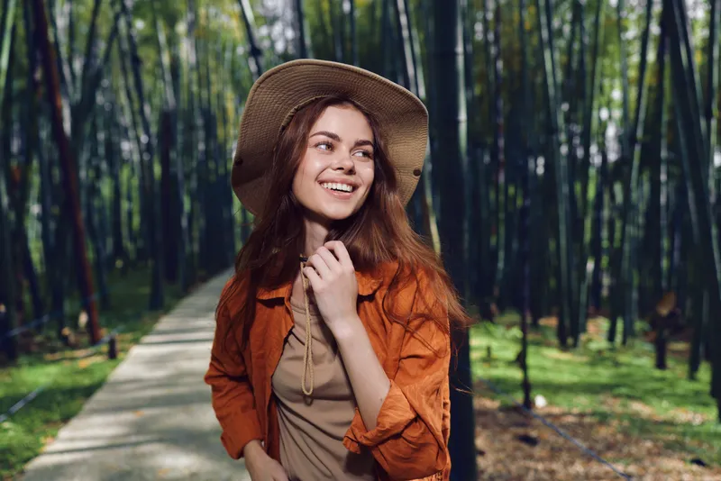 Woman with a warm smile wearing a straw hat walks along a forest path, portrait-style photo of a young brunette in casual jacket, sunlight and relaxed outdoor nature lifestyle.