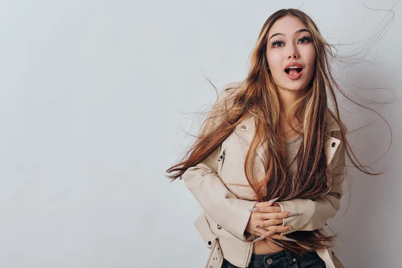 Young woman with long hair wearing beige jacket holding stomach with surprise or discomfort expression on light background for health or emotion concept.
